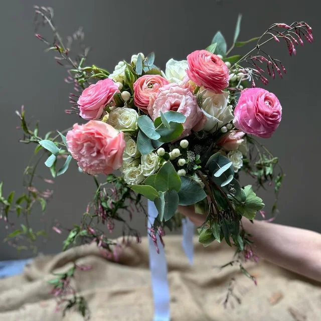 A hand-held bouquet of pink ranunculus, cream roses and flowing greenery