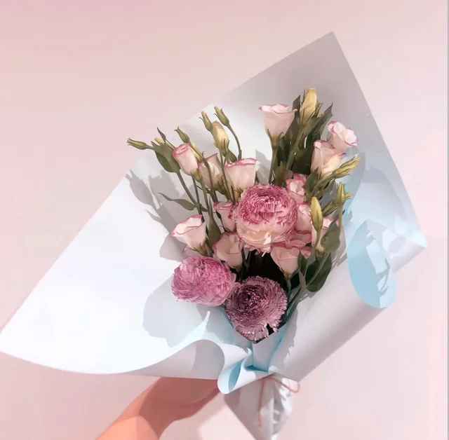 A small pink ranunculus and lisianthus bouquet in pale blue wrapping, held in the sunlight