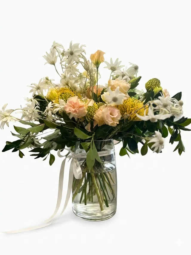 An airy yellow and cream flower arrangement in a clear glass vase with an Own Flower ribbon